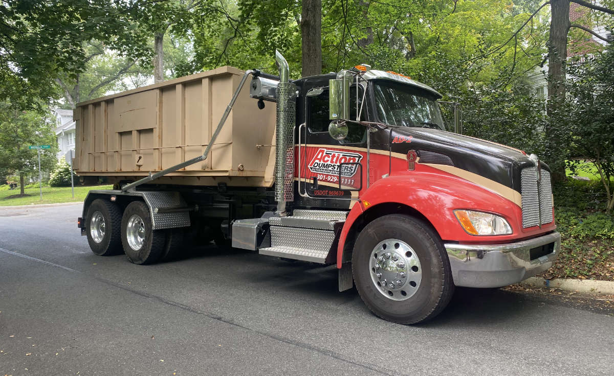 Action Dumpsters roll-off truck carrying a tan dumpster on a tree-lined residential street