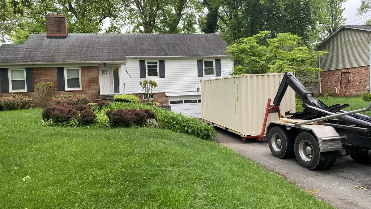 Roll-off truck setting a tan dumpster on a driveway in front of a brick and white house, serving Aspen Hill, MD
