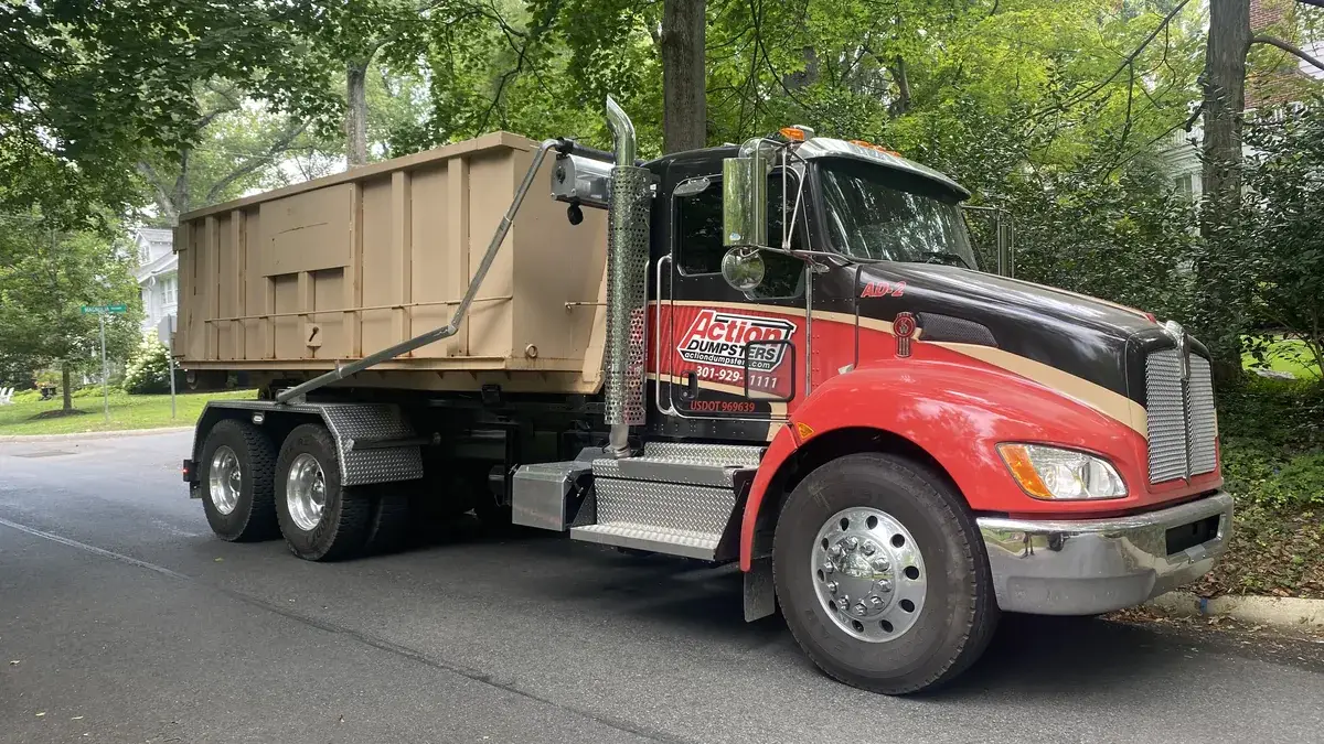 Red and black Action Dumpsters roll-off truck carrying a tan dumpster down a tree-lined street, serving Bethesda, MD