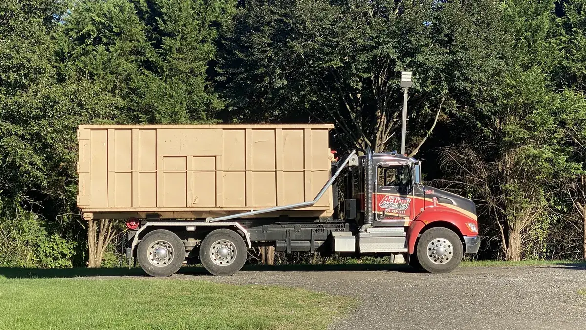 Action Dumpsters truck with a tan roll-off dumpster parked beside tall pines, serving Burtonsville, MD