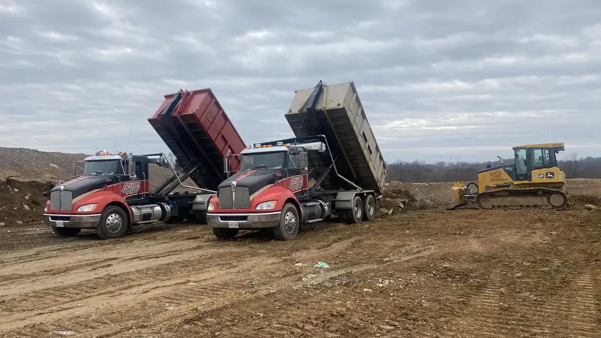Two Action Dumpsters trucks tipping out roll-off dumpsters beside a bulldozer at a dirt excavation site