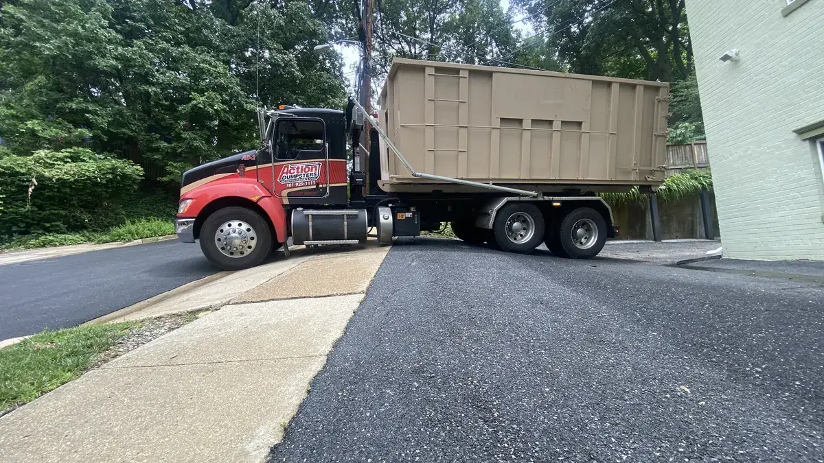 Action Dumpsters truck carrying a tan roll-off dumpster on a freshly paved lot, serving Silver Spring, MD